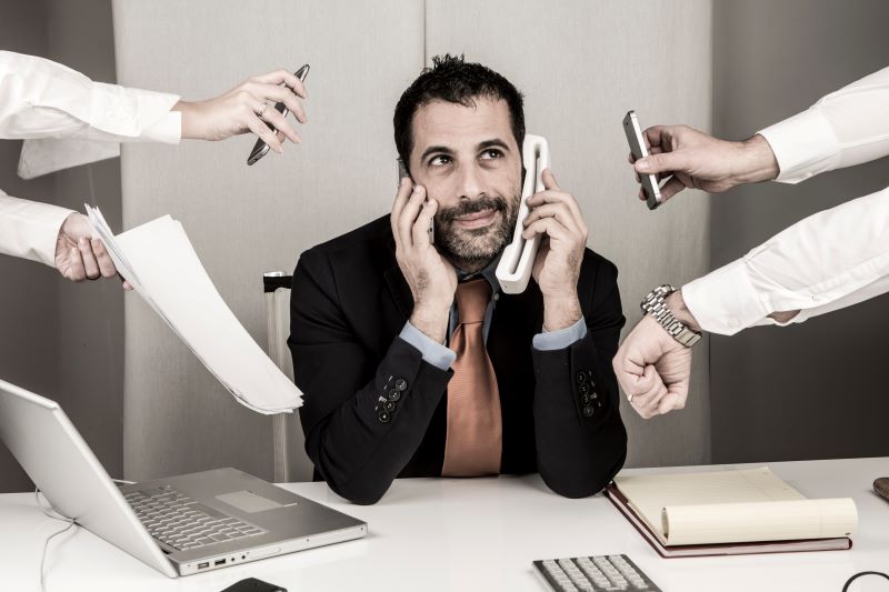 Homem atende telefones enquanto várias outras pessoas estendem as mãos lhe entregando outras tarefas. O acúmulo de funções é um dos gatilhos para o estresse ocupacional.