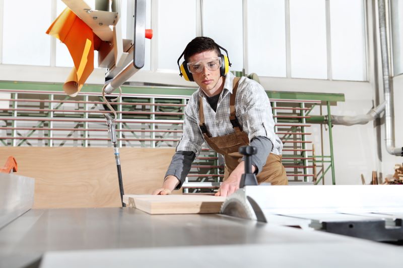 Homem carpinteiro usando serra circular de bancada enquanto usa abafadores nos ouvidos e óculos de proteção.