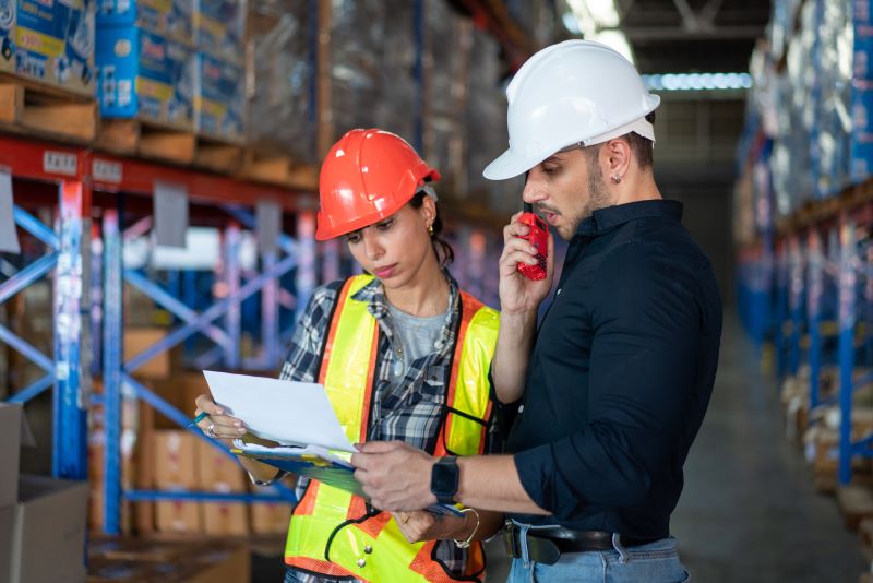 Engenheiro e engenheira analisam um documento de consultoria em segurança do trabalho num depósito de mercadorias.