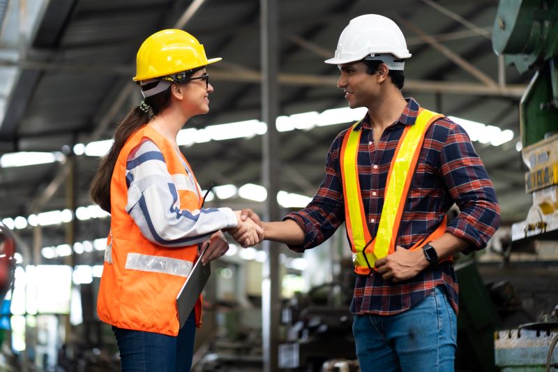 Homem e mulher vestindo EPIs apertam as mãos em indústria, simbolizando a contratação do serviço de segurança do trabalho.