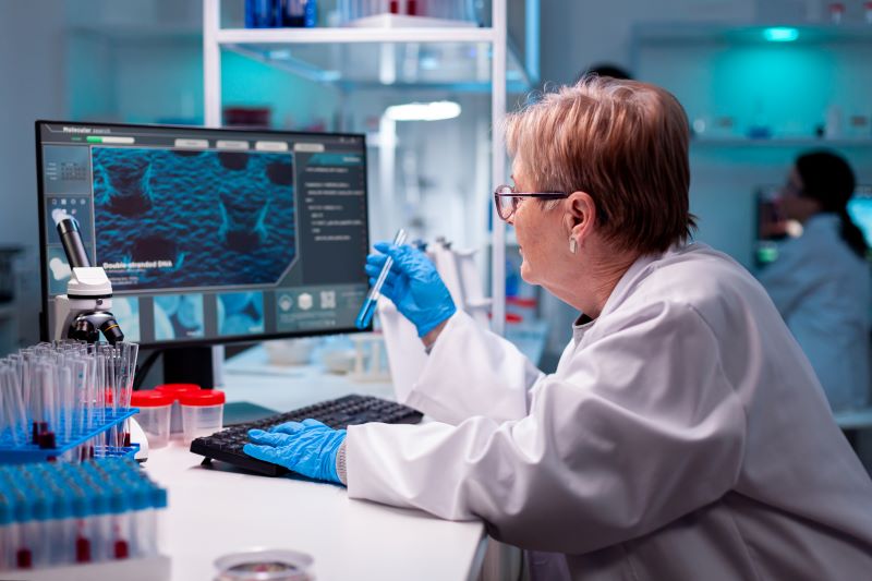 Em um laboratório, mulher de óculos, jaleco e luvas azuis segura um tubo de ensaio e olha para o computador que exibe diagnóstico.