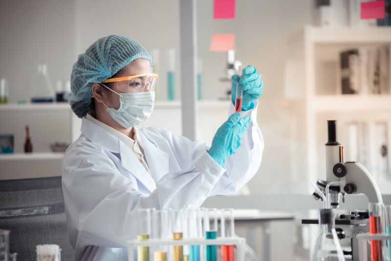 Uma cientista utilizando jaleco, luvas, óculos de proteção, gorro e máscara manuseia um tubo de ensaio em um laboratório.