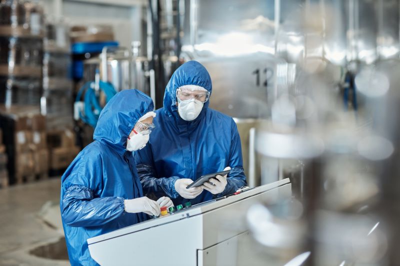 Dois trabalhadores em indústria química usando um tablet e equipados com macacão, luvas, óculos de proteção e máscaras.