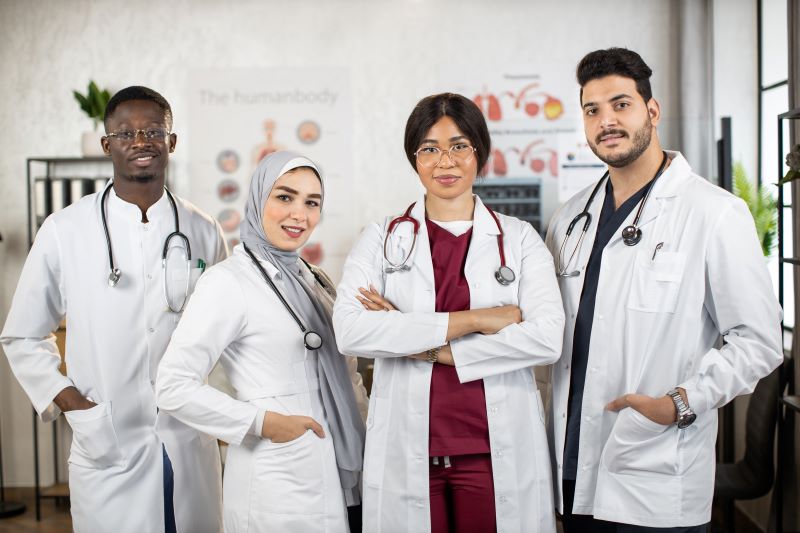 Quatro profissionais da saúde usando jalecos brancos e estetos pendurados no pescoço olham para frente, um ao lado do outro. 