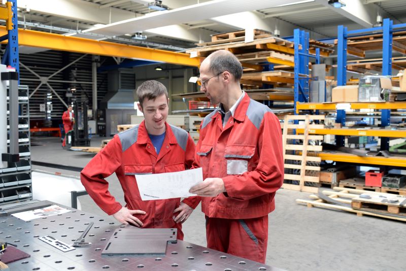 Em um galpão de fábrica, um homem usando uniforme mostra alguns papéis para outro, ensinando-o algo.