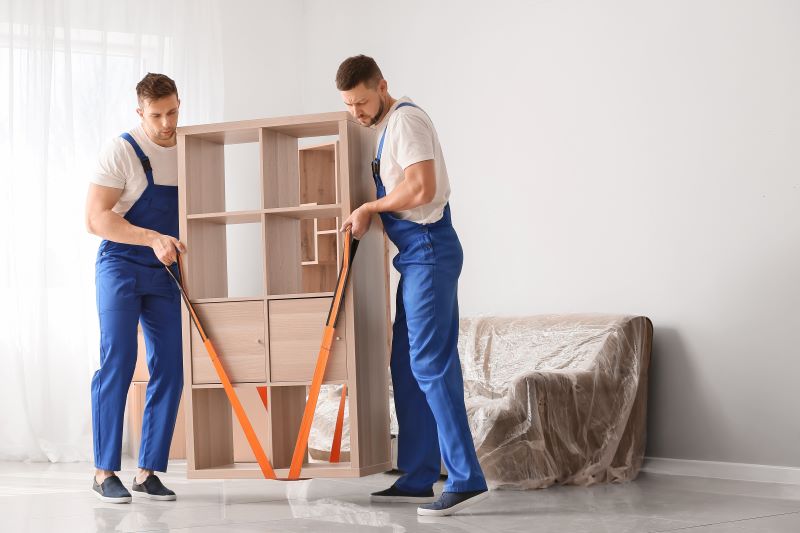 Dois trabalhadores utilizando cinta para o levantamento de carga, devidamente posicionando estante em sala de estar.