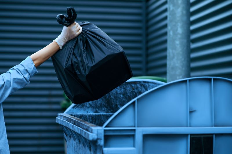 Gerenciamento de resíduos: funcionária jogando saco de lixo preto em lixeira comunitária nos fundos do estabelecimento.