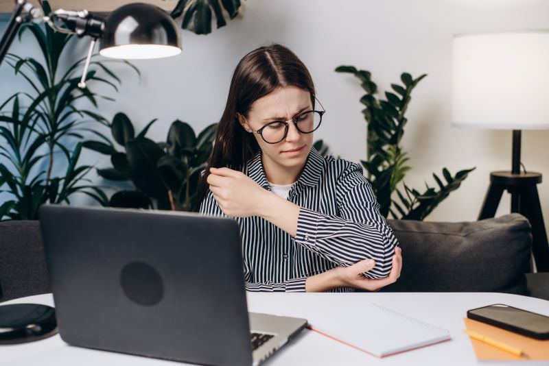 Mulher trabalhando no computador com lesão por esforço repetitivo no cotovelo.